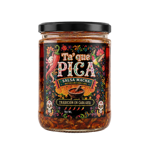 A glass jar of Ta' que Pica Salsa Macha Mexican Chili, featuring a colorful Day of the Dead style label with skulls, a snake, and a chili pepper, topped with a black lid on a white background.